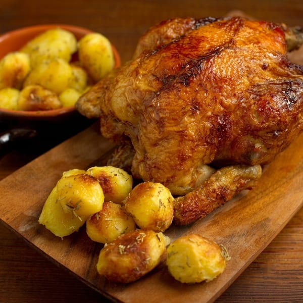 Whole roasted chicken with crispy skin, served on a wooden board with seasoned roasted potatoes.